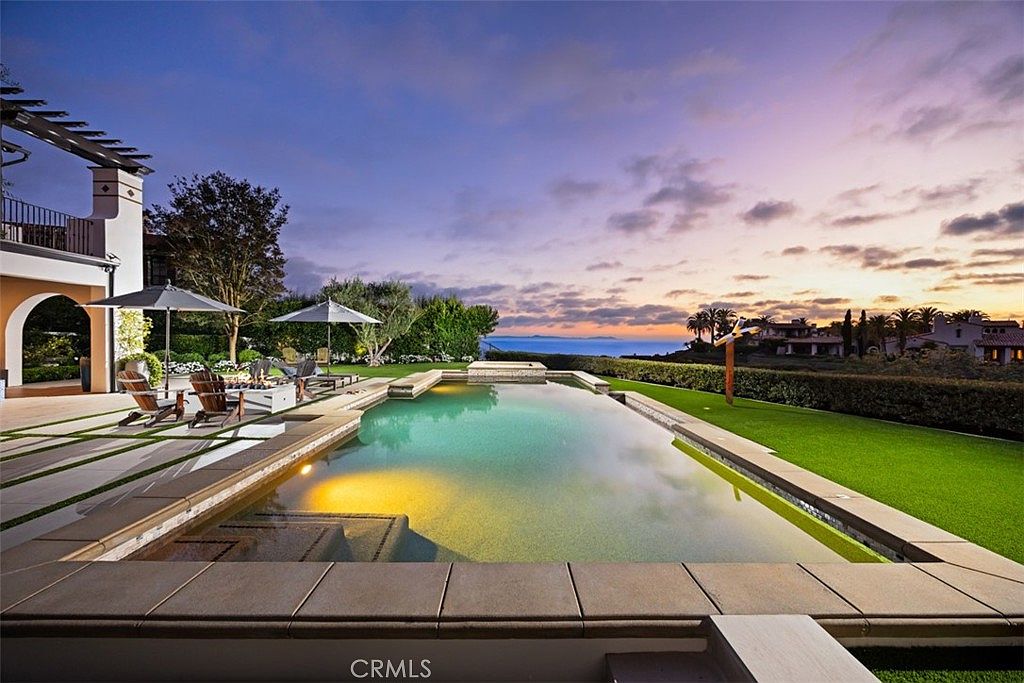 This stunning backyard features a rectangular infinity-edge pool with integrated lighting, set against a backdrop of a vibrant sunset over the ocean. A paved patio area with umbrellas and lounge chairs provides a comfortable space for outdoor relaxation, while a manicured lawn borders the pool deck. The scene captures a luxurious, serene atmosphere perfect for entertaining, with the horizon line emphasizing the property's elevated coastal position.