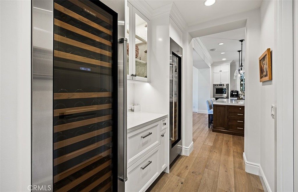 This image captures a sophisticated butler's pantry or wine storage area featuring a tall, glass-fronted wine refrigerator with wooden shelving. The space includes white cabinetry with a light-colored countertop and glass-fronted upper cabinets, leading into a bright, open-concept kitchen with light wood flooring. The perspective is a straight-on view through a transition space, highlighting the clean, high-end finishes and seamless flow between rooms.