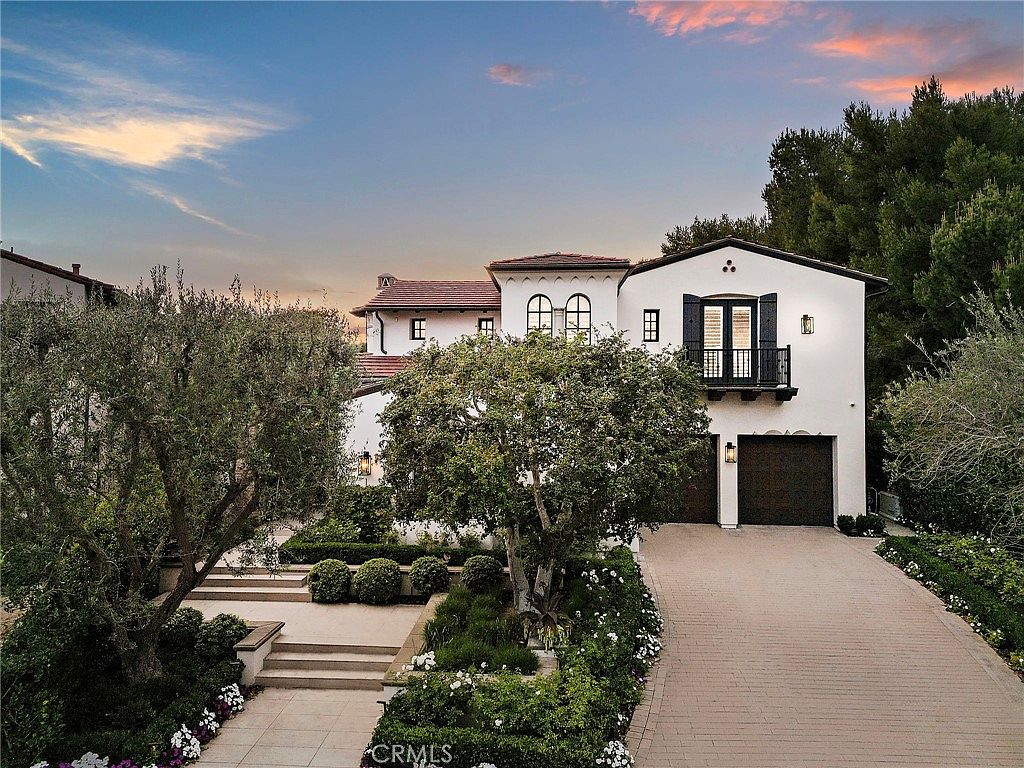 This elegant Mediterranean-style residence features a crisp white stucco facade, a terracotta tile roof, and a prominent arched balcony with black iron railings. The front exterior is complemented by a paved driveway leading to a two-car garage, lush manicured landscaping, and a tiered stone staircase leading to the entrance. The perspective is a wide-angle shot captured during the golden hour, highlighting the warm, inviting atmosphere of the property.