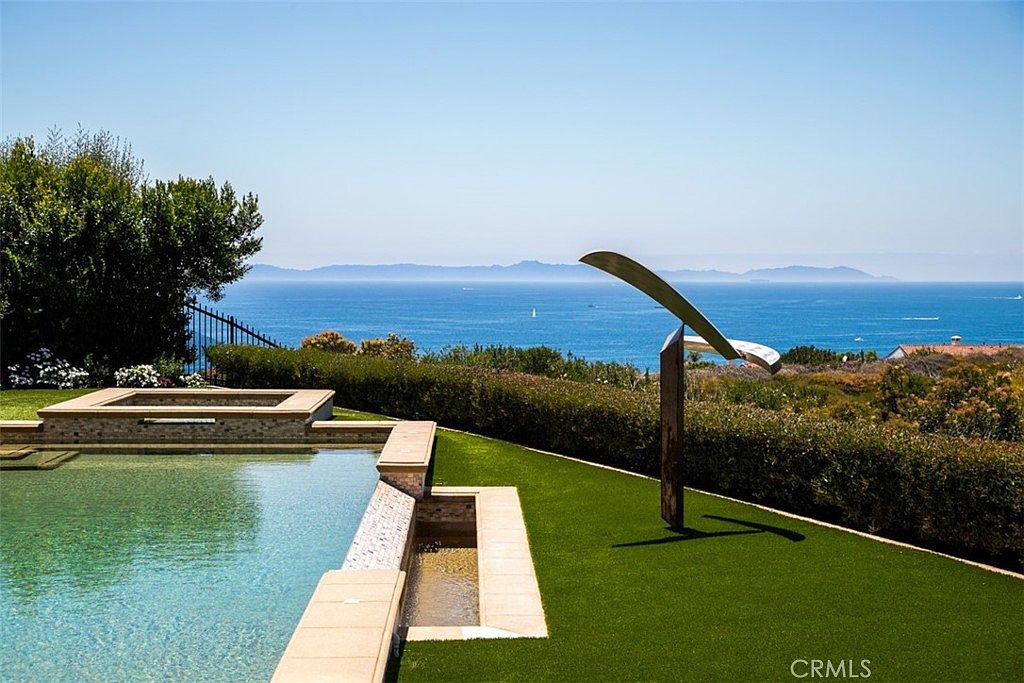 This outdoor space features a sleek, modern swimming pool with a stone-tiled edge and a raised spa section. A manicured green lawn stretches toward a low hedge, which frames a breathtaking, unobstructed panoramic view of the deep blue ocean and distant mountains. A unique, abstract metal sculpture stands prominently on the grass, adding a contemporary artistic element to the serene coastal landscape.