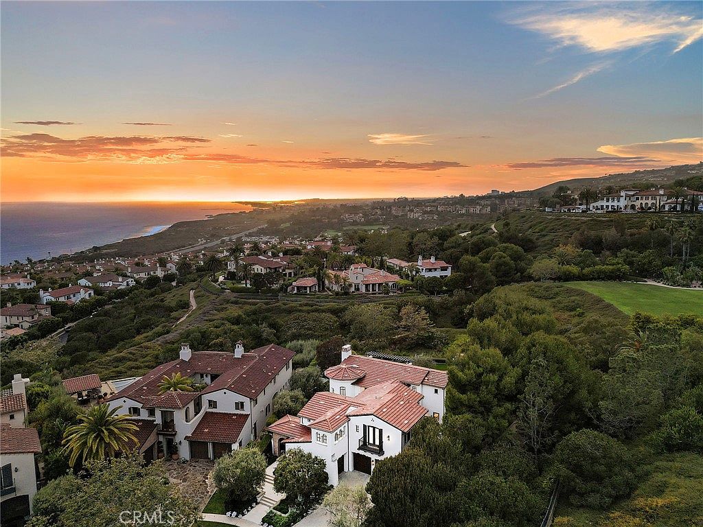 This high-angle aerial shot captures a sprawling Mediterranean-style estate nestled within a lush, coastal landscape at sunset. The property features multiple white-stucco buildings with terracotta-tiled roofs, surrounded by mature trees and manicured greenery, with the vast Pacific Ocean visible in the distance. The perspective emphasizes the home's prime location, showcasing the expansive coastal views and the serene, upscale neighborhood setting.