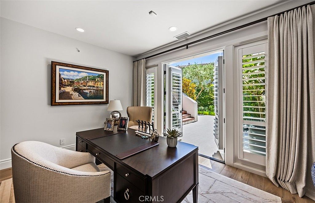 This elegant home office features a dark wood desk centered in the room, paired with a light-colored upholstered chair. Large glass doors with white shutters open to a sunlit outdoor patio, creating a seamless transition between indoor and outdoor spaces. The room is decorated with a framed landscape painting and soft, neutral-toned curtains, offering a sophisticated and tranquil workspace.