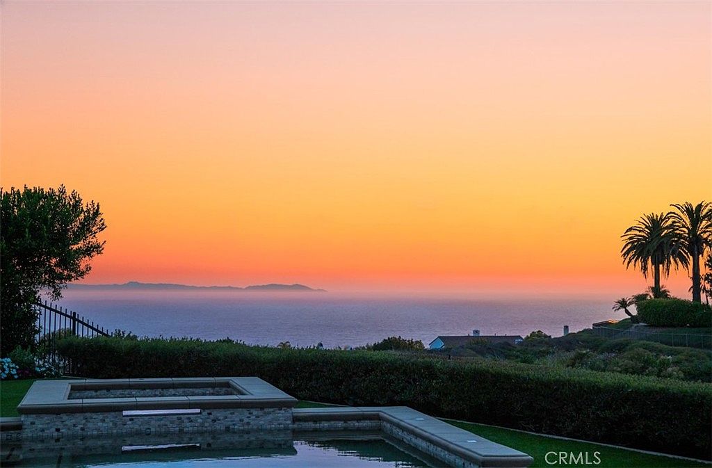 This image captures a serene sunset view from a backyard pool area, overlooking a vast ocean horizon with distant islands silhouetted against a vibrant orange and pink sky. In the foreground, the edge of a stone-tiled spa or pool is visible, framed by manicured hedges and lush landscaping. The perspective is low and wide, emphasizing the tranquil atmosphere and the expansive, picturesque coastal vista.