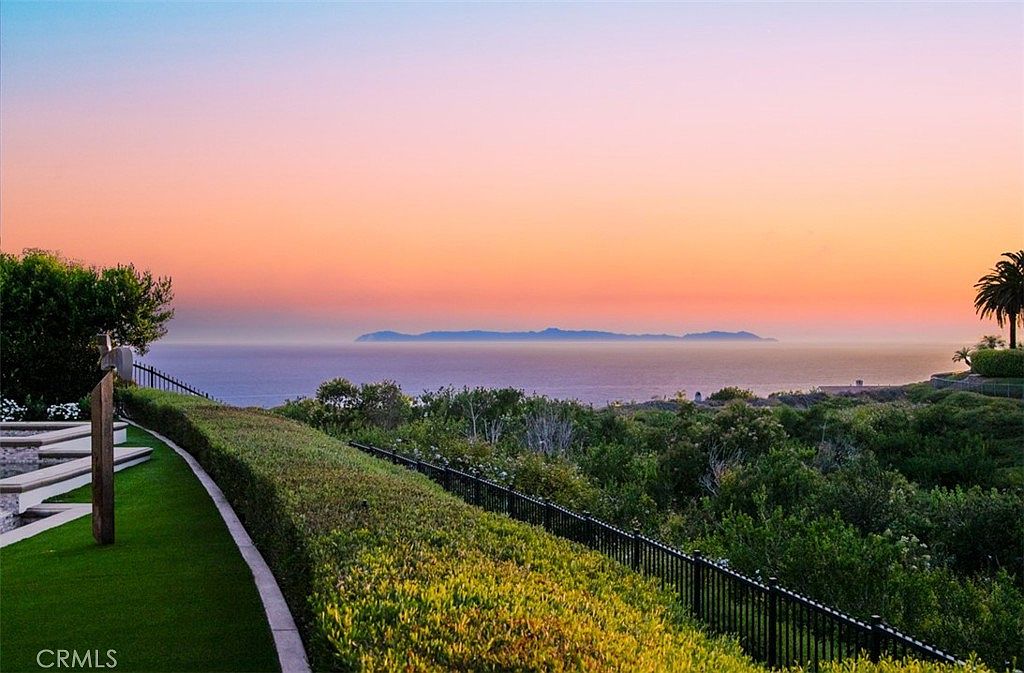 This scenic outdoor space features a manicured lawn bordered by a neatly trimmed hedge and a black metal fence, overlooking a vast ocean horizon. In the distance, a prominent island silhouette is visible against a vibrant, multi-colored sunset sky. The perspective is elevated, capturing the serene transition from the lush foreground greenery to the expansive coastal view.
