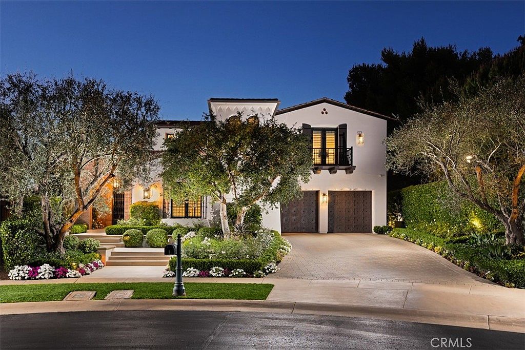 This elegant Mediterranean-style residence is captured at dusk, showcasing a white stucco facade with dark wood-toned garage doors and a matching balcony railing. The front yard is meticulously landscaped with manicured hedges, mature trees, and a stone-paved driveway leading to the entrance. Warm exterior lighting highlights the architectural details, creating an inviting and sophisticated curb appeal.