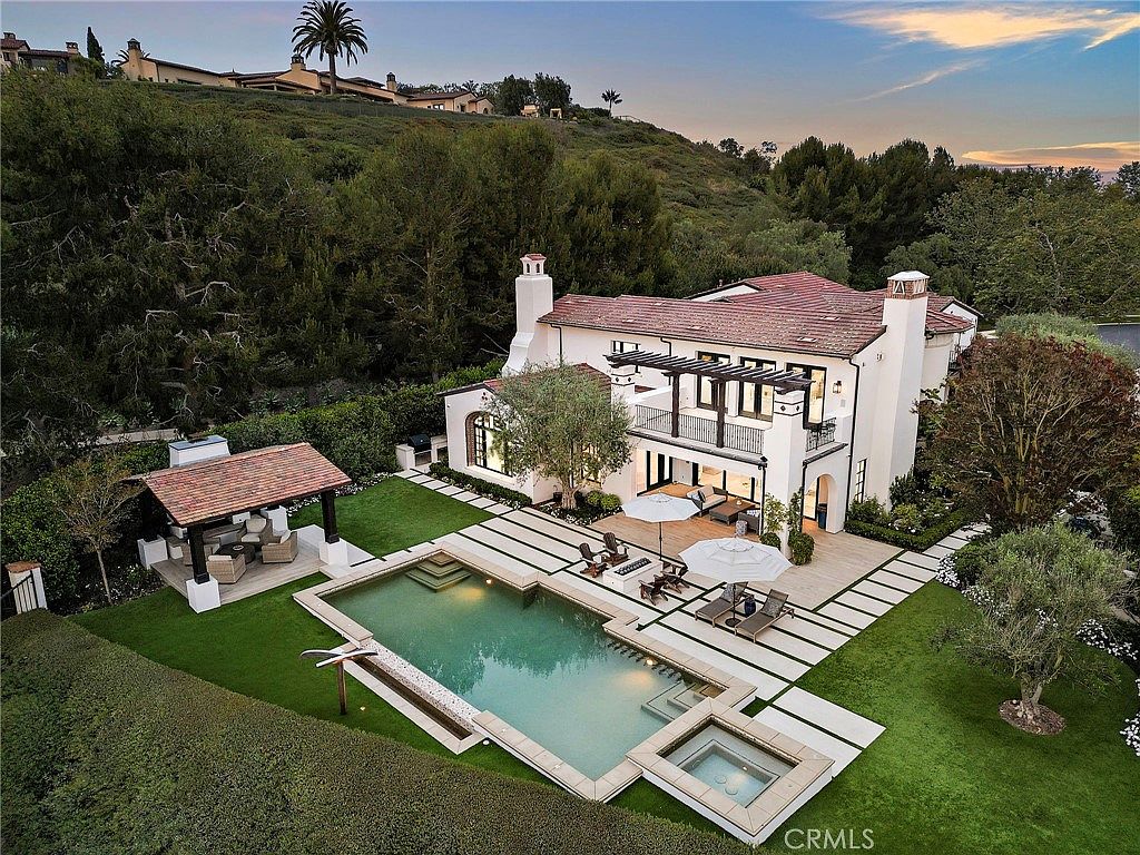 This elevated aerial perspective showcases a luxurious Mediterranean-style estate featuring a sprawling backyard with a custom swimming pool and integrated spa. The property includes a covered outdoor pavilion with a fireplace, manicured green lawns, and a multi-level terrace with a balcony overlooking the pool area. The home is nestled against a lush, tree-lined hillside, creating a private and serene atmosphere.