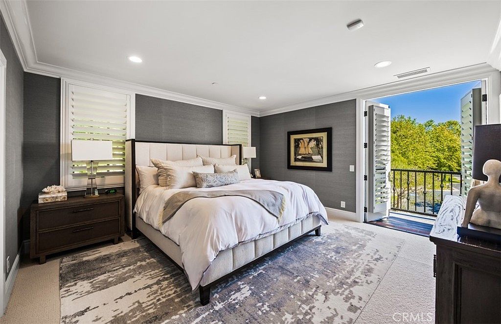 This elegant primary bedroom features a plush king-sized bed with a tufted headboard, centered against a dark, textured accent wall. The room is anchored by a large, abstract-patterned area rug and includes a dark wood nightstand and a matching dresser. A set of open French doors leads to a private balcony, allowing natural light to flood the space and providing a scenic view of the surrounding trees.