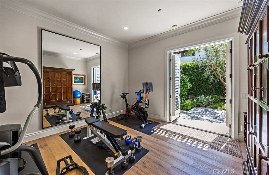 This well-appointed home gym features light wood flooring, a large wall-mounted mirror, and a variety of fitness equipment including a stationary bike and a weight bench. The room opens directly to a lush outdoor garden through wide double doors, creating a bright and airy atmosphere. The space is designed for convenience and wellness, blending indoor exercise functionality with a seamless connection to nature.