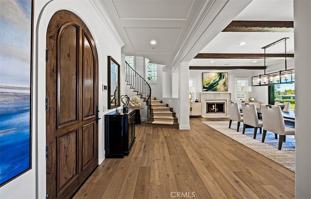 This elegant entryway features a grand arched wooden front door that opens into a spacious foyer with light wood flooring. A curved staircase with a decorative iron railing leads to the upper level, while the open-concept layout flows seamlessly into a dining area accented by exposed ceiling beams and a fireplace with a stone surround. The space is bright and airy, showcasing a sophisticated design that blends traditional architectural elements with a modern, open floor plan.