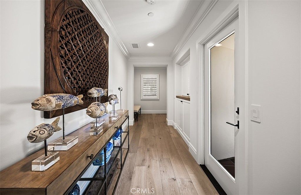 This bright, modern hallway features light wood flooring and crisp white walls, creating an airy and welcoming atmosphere. A long console table adorned with decorative fish sculptures and a large, intricate circular wall art piece serves as a focal point along the left wall. The perspective leads the eye down the corridor toward a secondary space with a bench, framed by a glass-paneled door on the right.