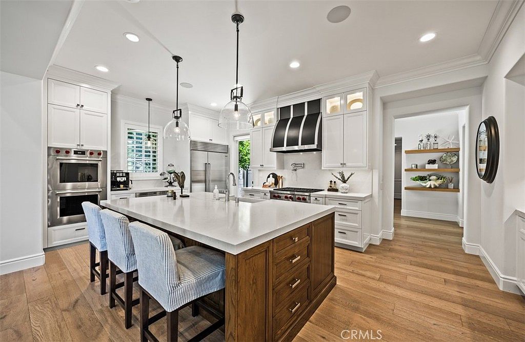This bright and spacious kitchen features a large central island with a light-colored countertop and wood-toned cabinetry, complemented by white perimeter cabinets and stainless steel appliances. The room is illuminated by elegant pendant lights and recessed lighting, creating an inviting atmosphere. A wide doorway leads to a hallway with open shelving, while light-toned wood flooring runs throughout the space.