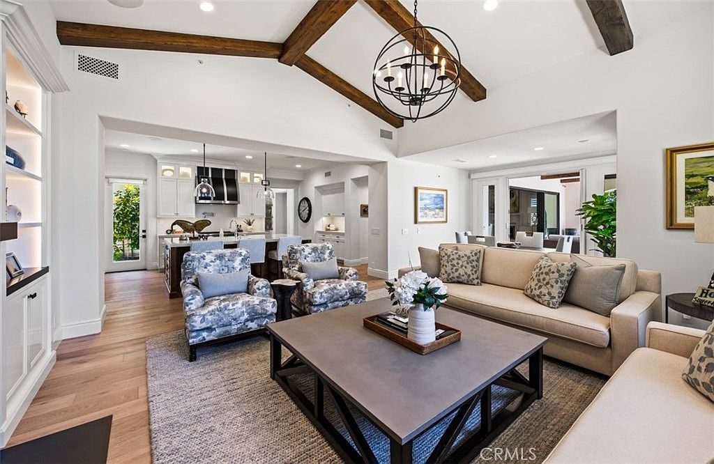 This spacious, open-concept living area features vaulted ceilings with exposed wood beams and a large, dark-toned chandelier as a central focal point. The room is furnished with a neutral-toned sofa, two patterned armchairs, and a substantial square coffee table, all set upon a textured area rug. In the background, a bright, modern kitchen with a large island and white cabinetry is visible, creating a seamless flow between the living and cooking spaces.