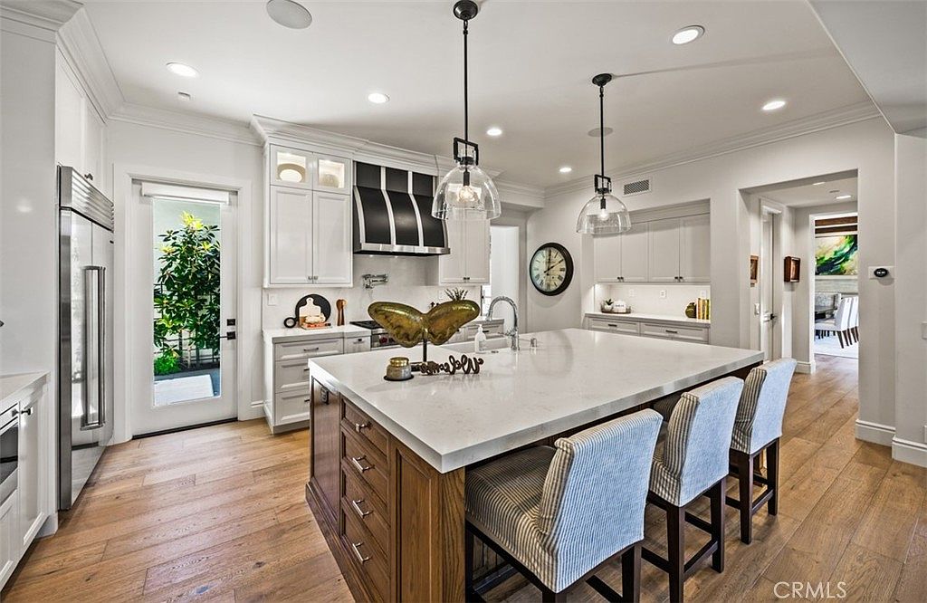 This bright and spacious kitchen features a large central island with a light-toned stone countertop and wood cabinetry, providing ample seating with three upholstered stools. The perimeter includes white cabinetry, a stainless steel refrigerator, and a decorative range hood, all illuminated by recessed lighting and two glass pendant lights. The perspective captures the open-concept layout, highlighting the light wood flooring and a glass door that leads to an outdoor space.