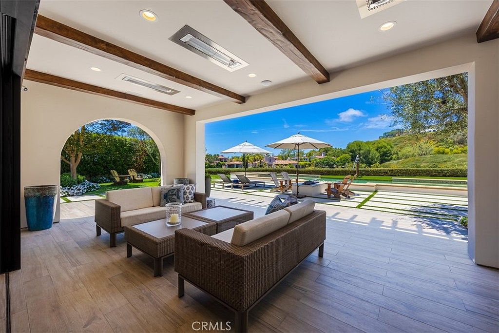This expansive covered patio offers a seamless transition to the outdoors, featuring a comfortable seating area with modern wicker furniture and a neutral color palette. The space is defined by exposed wood ceiling beams and recessed lighting, overlooking a pristine swimming pool and a lush, landscaped hillside. The open-air design creates a luxurious, resort-like atmosphere perfect for entertaining.