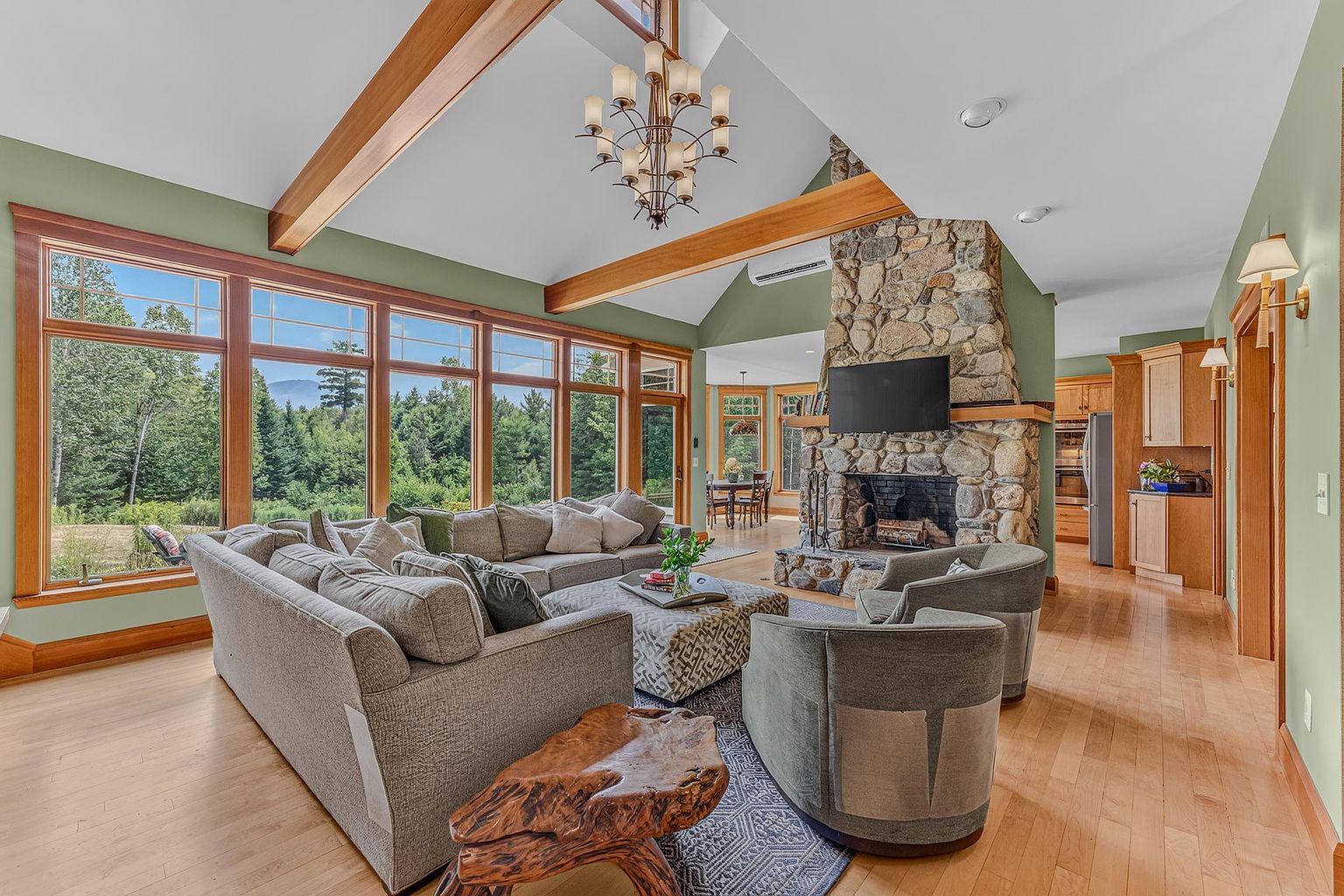This spacious living room features a vaulted ceiling with exposed wooden beams and a large stone-clad chimney breast that serves as a focal point. A large sectional sofa and two armchairs are arranged around a patterned ottoman, all resting on a textured area rug atop light wood flooring. Expansive windows along the left wall provide abundant natural light and scenic views of the surrounding forest, creating an airy and inviting atmosphere.