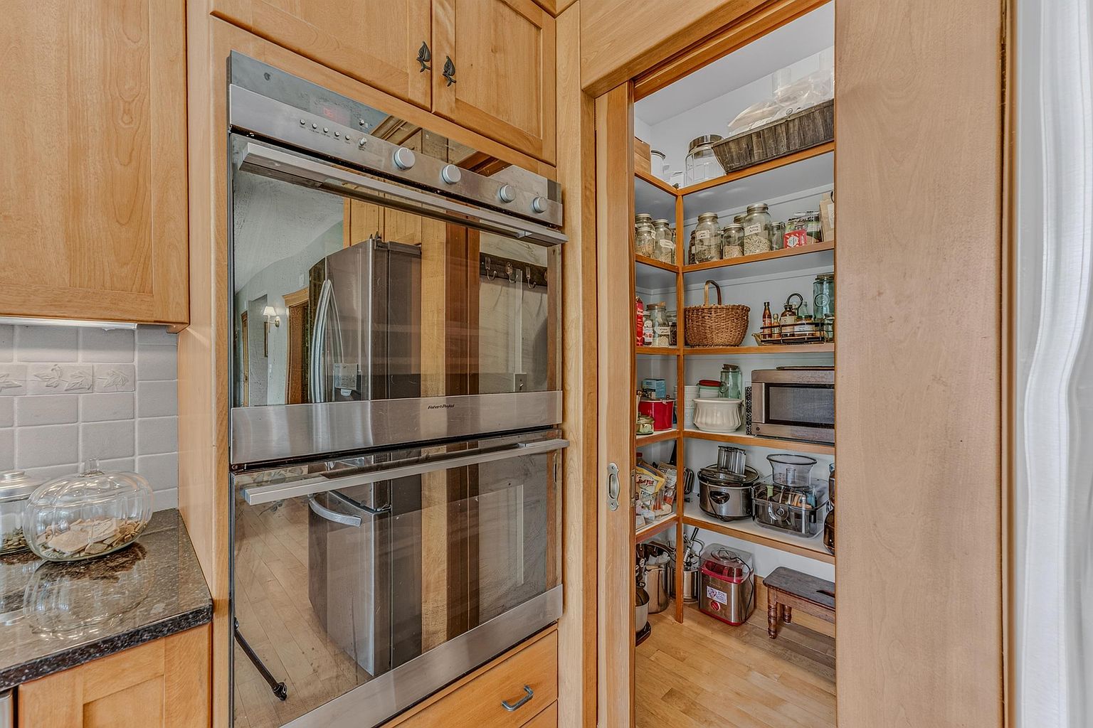 This kitchen view highlights a built-in stainless steel double oven integrated into light wood cabinetry, positioned next to a sliding door that reveals a well-organized walk-in pantry. The pantry features multiple shelves stocked with various jars, baskets, and small kitchen appliances, showcasing efficient storage solutions. A dark stone countertop with a glass jar sits in the foreground, contributing to the functional and warm aesthetic of the space.