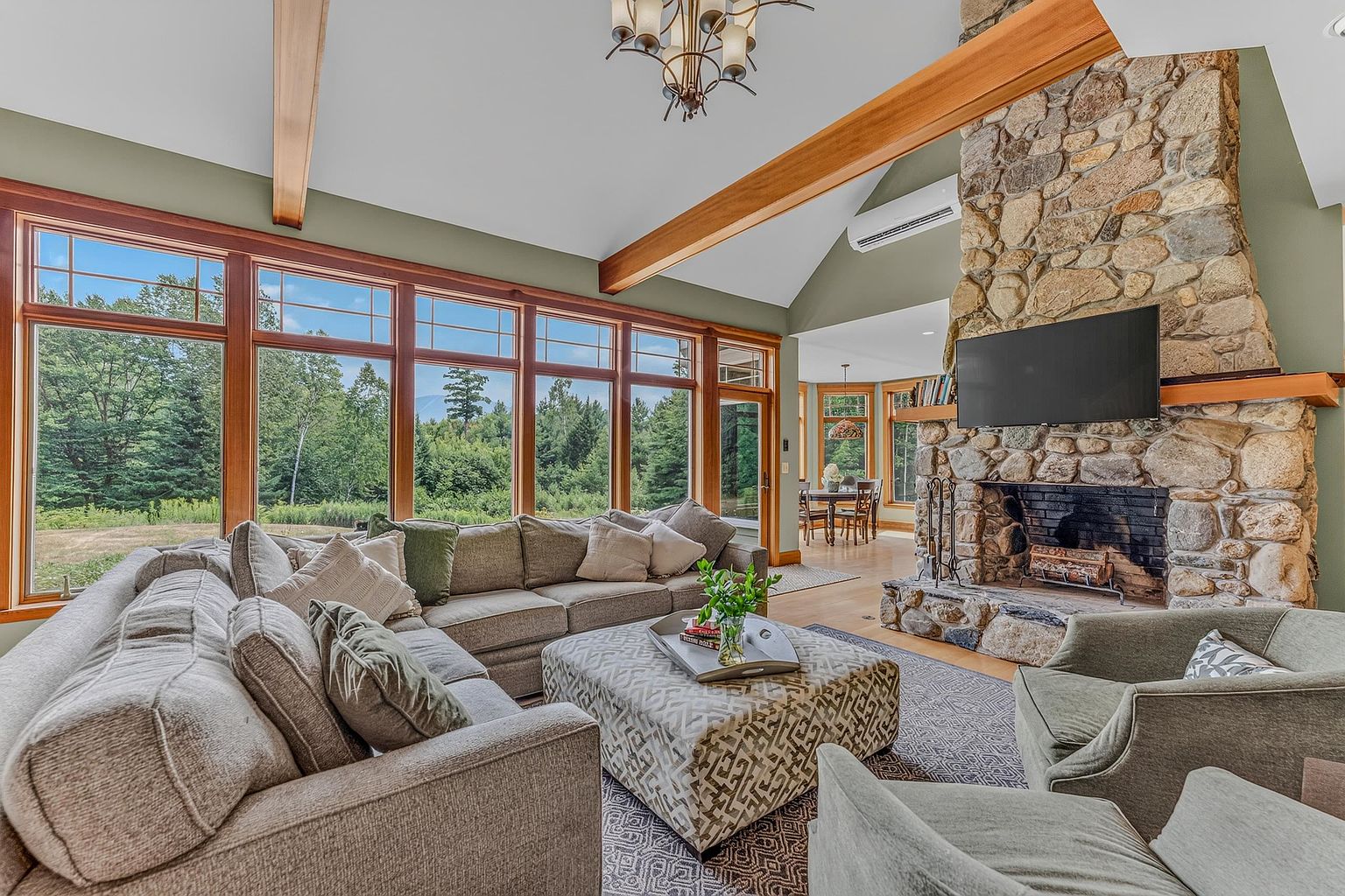 This spacious living room features a vaulted ceiling with exposed wood beams and a prominent stone-clad chimney breast housing a fireplace. A large sectional sofa and patterned ottoman are arranged on a textured rug, while a wall of expansive windows provides abundant natural light and views of the surrounding landscape. The open-concept design flows into a dining area, creating an inviting and airy atmosphere suitable for entertaining.