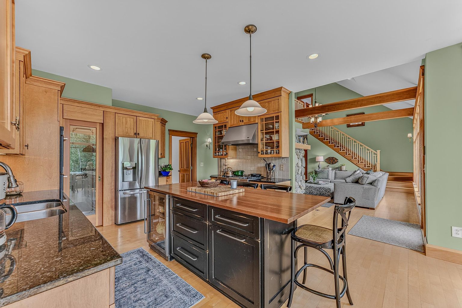 This open-concept kitchen features a large central island with a dark base and a light wood countertop, complemented by light wood cabinetry and dark stone countertops. The space flows seamlessly into a living area with a staircase and a stone-clad chimney breast, creating a warm and inviting atmosphere. The perspective is from the kitchen looking toward the living room, highlighting the cohesive design and natural light throughout the home.