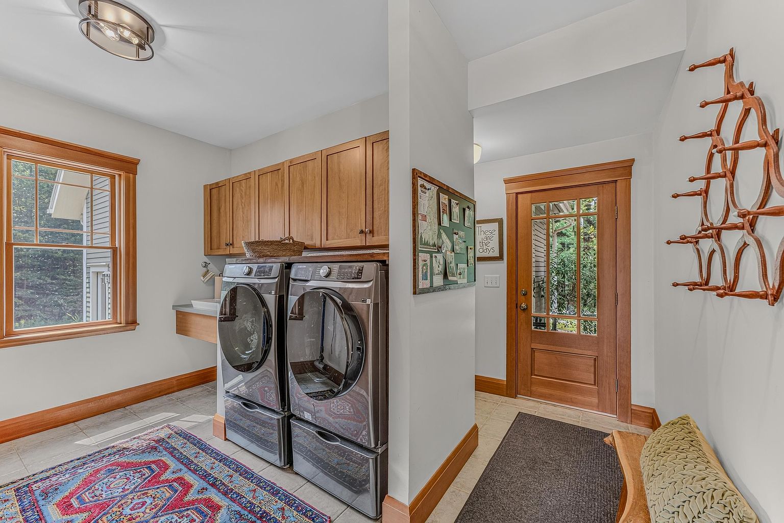 This functional laundry room features a pair of dark metallic front-loading washer and dryer units positioned beneath light wood cabinetry. A patterned area rug adds color to the tiled floor, while a wooden door with glass panes provides natural light and access to the outdoors. To the right, a wall-mounted wooden coat rack and a small bench with a textured cushion create a convenient mudroom-style entry area.