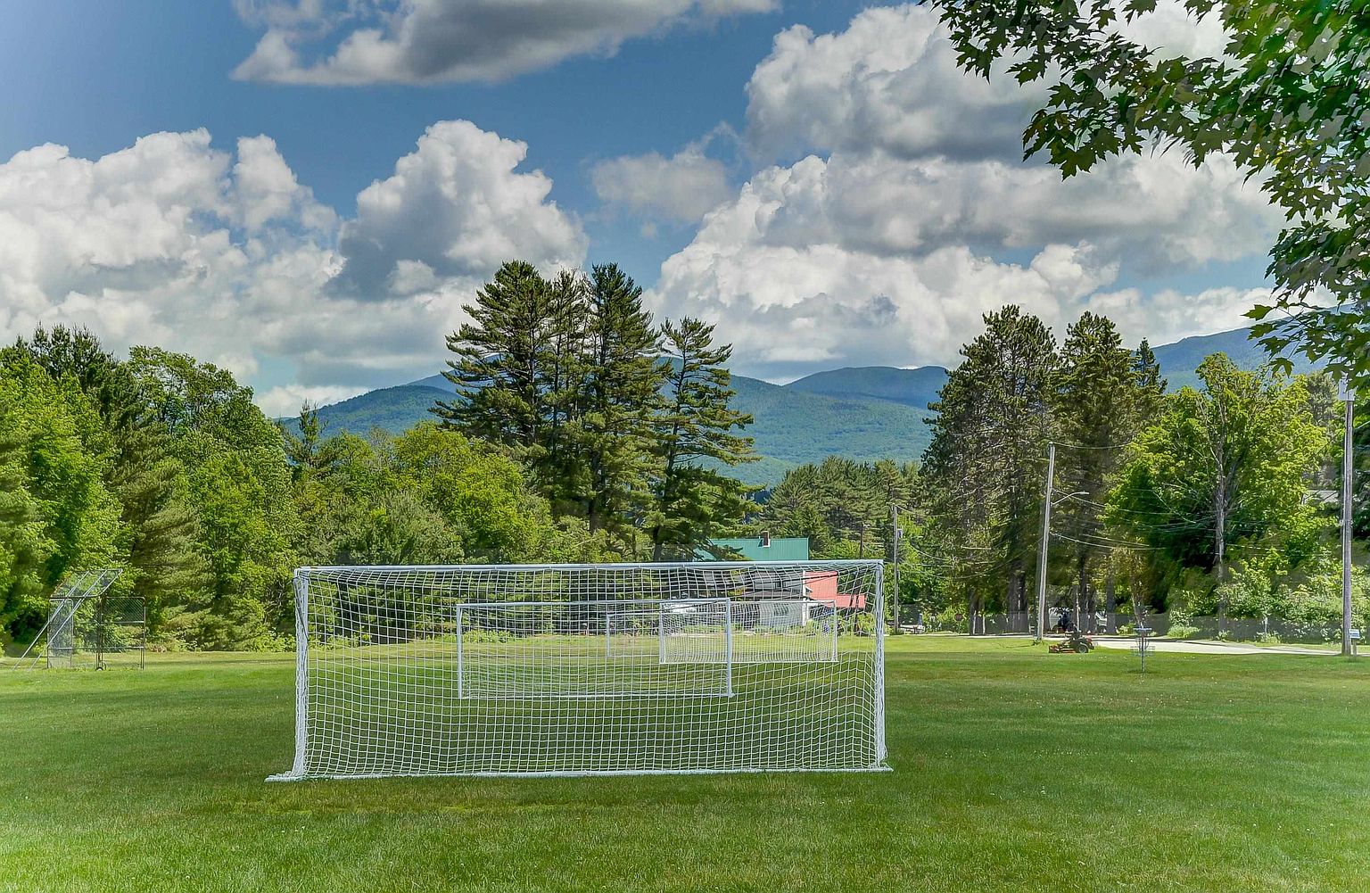 This image captures a wide, open grassy field featuring a soccer goal in the foreground, set against a backdrop of lush green trees and rolling mountains under a bright, cloudy sky. The perspective is at eye level, emphasizing the expansive recreational space available for outdoor activities. The scene conveys a peaceful, scenic environment typical of a community park or large residential property grounds.