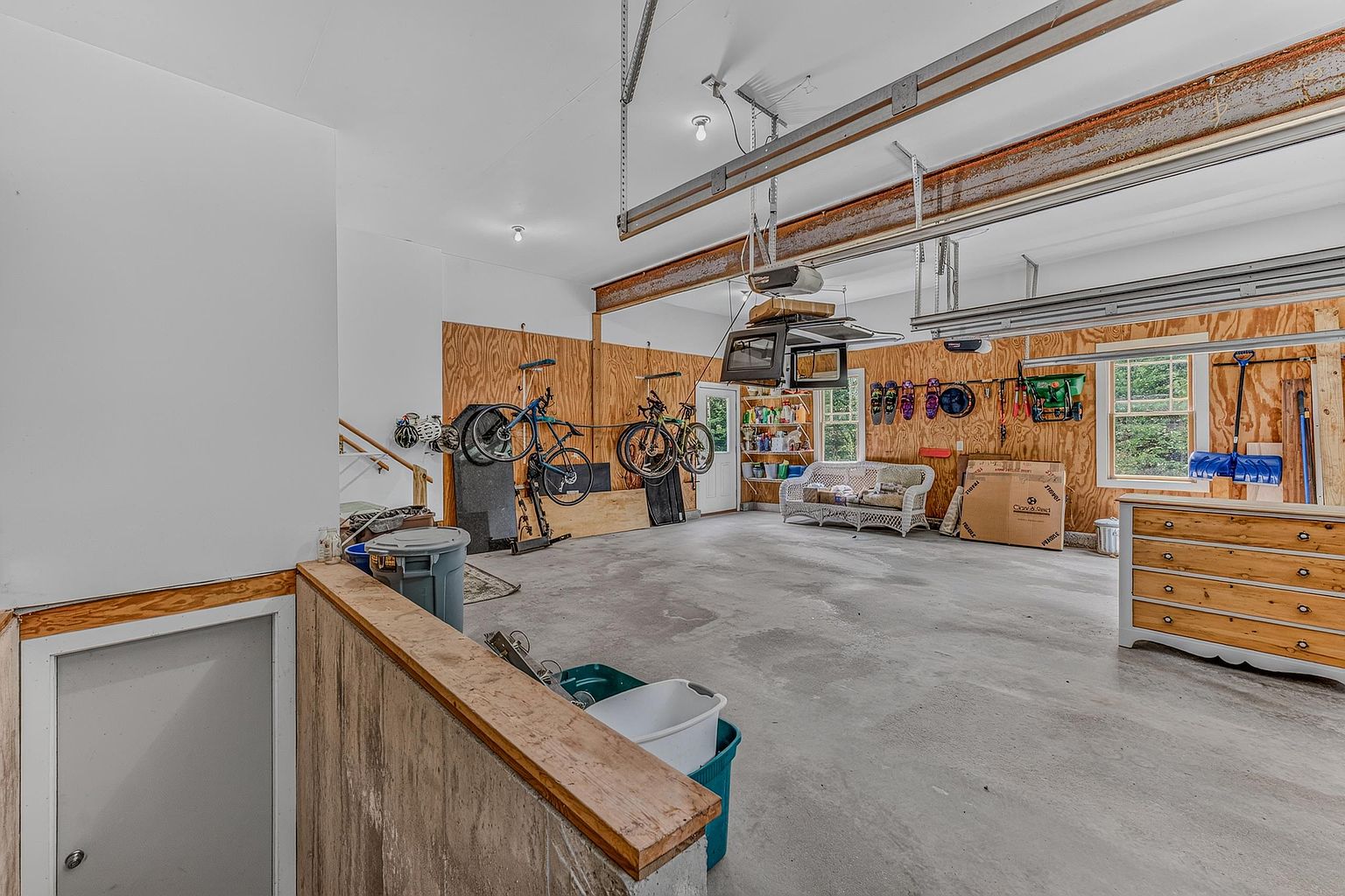 This spacious, unfinished garage features exposed plywood walls and a concrete floor, offering ample room for storage or workshop activities. The space includes a white wicker sofa, two bicycles mounted on the wall, and a wooden dresser, suggesting a multi-purpose utility area. A set of stairs leads down to a lower level, while a window provides natural light to the interior.