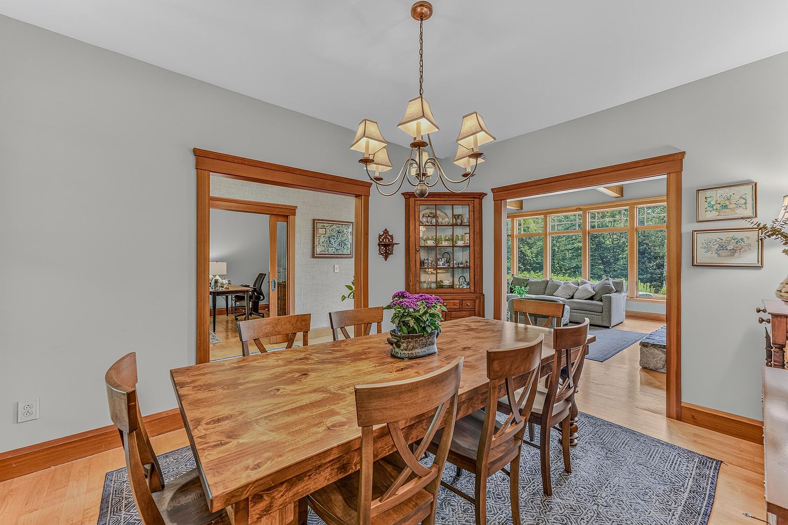 This dining room features a large, rustic wood table as the centerpiece, surrounded by matching chairs and illuminated by a traditional multi-arm chandelier. The space is framed by warm wood trim around the doorways, which lead into an adjacent office and a bright living area with large windows. A corner china cabinet adds a classic touch, while the neutral wall color and light wood flooring create an inviting and cohesive atmosphere.