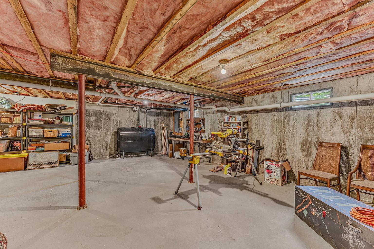 This unfinished basement features exposed ceiling joists with pink insulation, concrete walls, and a smooth concrete floor. The space is supported by two prominent red metal support columns and includes a large black oil tank, a workbench area with power tools, and storage shelving. The perspective is a wide-angle view from the center of the room, highlighting the utility and workshop potential of the unfinished lower level.