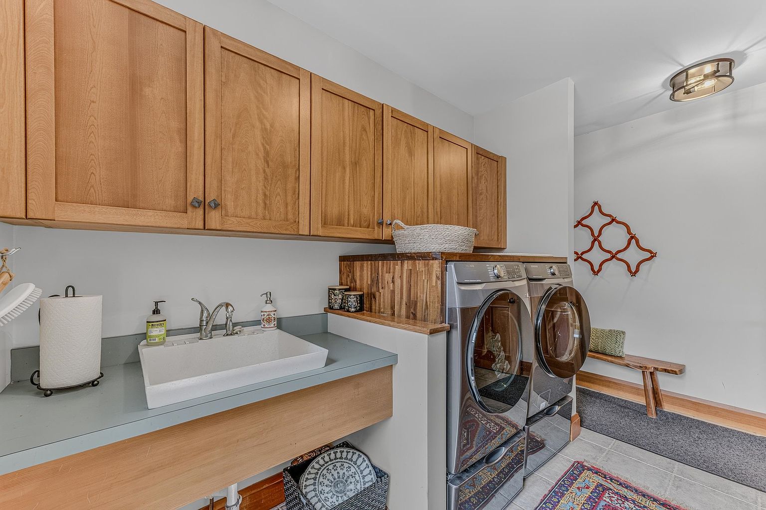 This functional laundry room features a utility sink set into a gray countertop with light wood cabinetry above for storage. A side-by-side washer and dryer set is positioned next to a custom wood-topped counter, creating a cohesive workspace. The room includes a small wooden bench and a decorative wall accent, providing a practical yet inviting space for household chores.