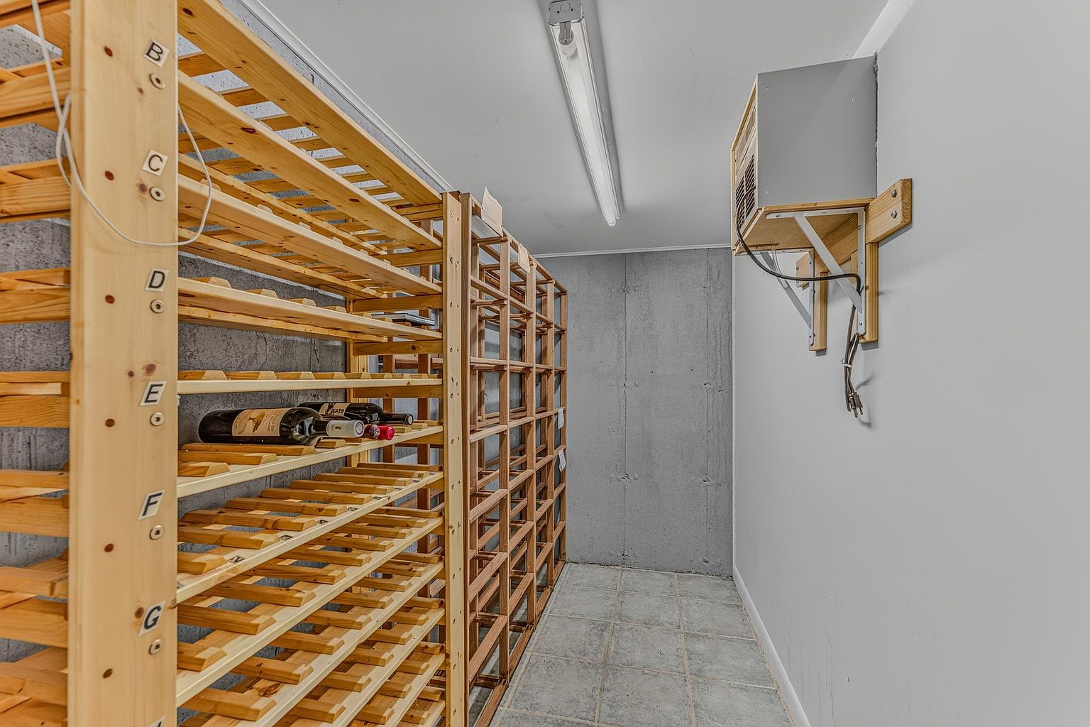 This dedicated wine cellar features custom light wood racking systems with labeled shelving units for organized storage. The space is finished with a concrete wall and tiled flooring, creating a functional and utilitarian aesthetic. A wall-mounted cooling unit is positioned to maintain the climate, and the perspective looks down the narrow aisle between the storage racks.