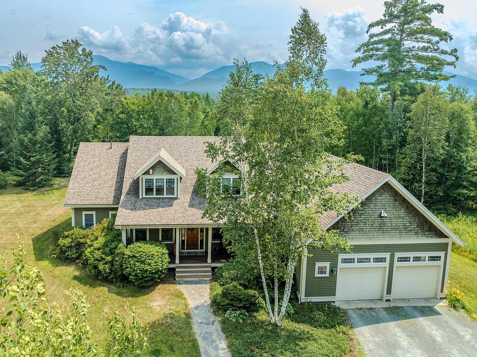 This elevated perspective captures a charming, green-sided home nestled in a lush, wooded landscape with a stunning mountain backdrop. The house features a multi-gabled roofline, a stone walkway leading to the front entrance, and an attached two-car garage. A prominent birch tree stands in the front yard, adding to the serene and private atmosphere of this mountain-style residence.