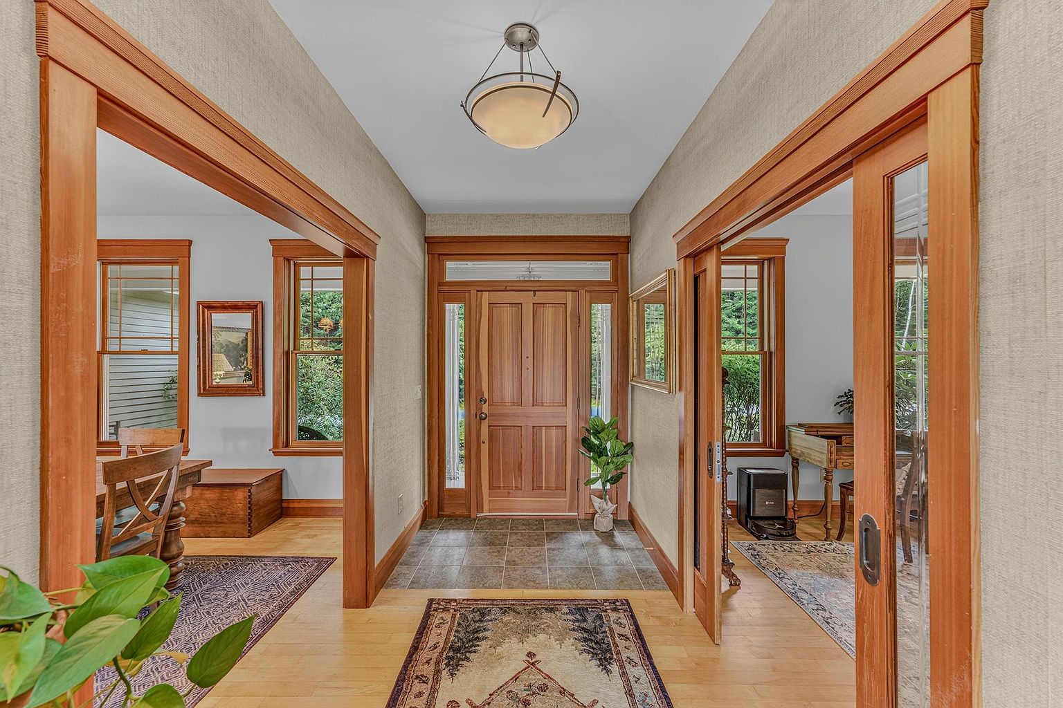 This inviting entryway features warm wood trim and light-toned wood flooring, creating a cohesive and traditional aesthetic. The space is framed by wide doorways leading into adjacent rooms, with a central front door flanked by sidelights that allow natural light to filter in. A decorative ceiling fixture hangs above, while patterned area rugs add texture and color to the transition area.