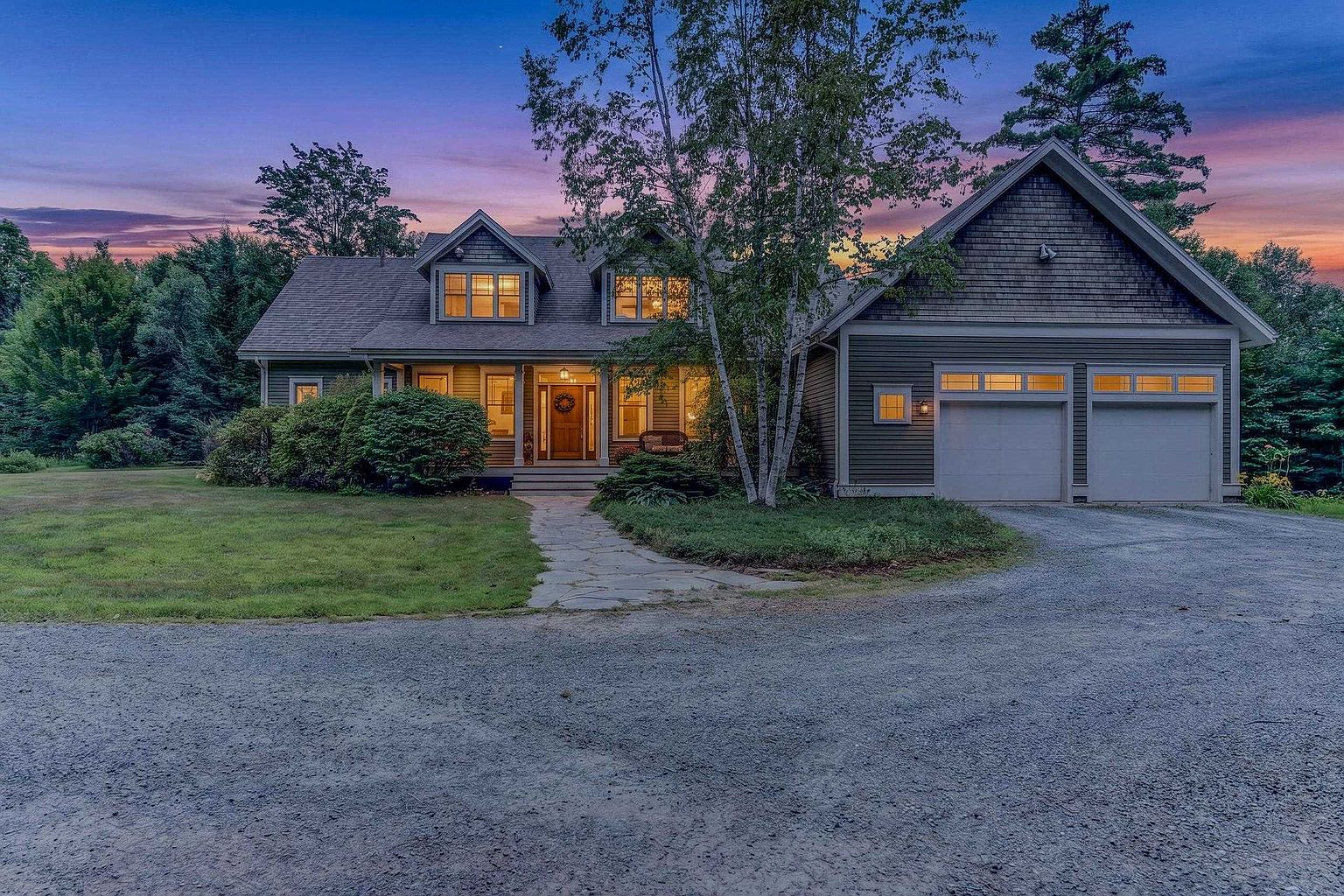 This charming two-story home features a classic shingle-style exterior with a welcoming front entryway and a two-car attached garage. The house is set against a vibrant sunset sky, with a stone walkway leading to the front door and a well-maintained lawn in the foreground. The architectural design includes multiple gables and dormer windows, creating a warm and inviting curb appeal.