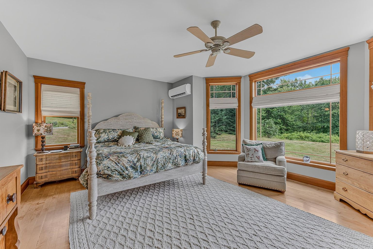 This spacious primary bedroom features light-toned hardwood flooring and a neutral color palette, centered around a large bed with a decorative headboard. The room is bathed in natural light from multiple large windows that offer a serene view of the surrounding greenery. A comfortable armchair sits by the window, and the space is accented by light wood furniture and a textured area rug, creating a peaceful and inviting atmosphere.