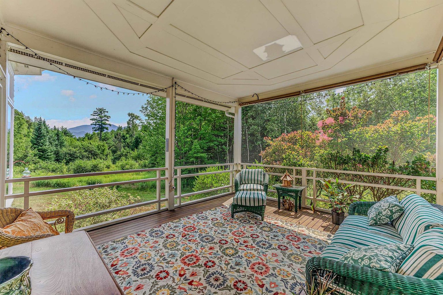 This inviting screened-in porch offers a serene outdoor living space with a view of lush green trees and distant mountains. The area is furnished with a striped wicker sofa and matching armchair, centered around a colorful floral area rug on the wooden deck. The space is framed by white railings and a high, paneled ceiling, creating a peaceful retreat perfect for relaxing or entertaining.