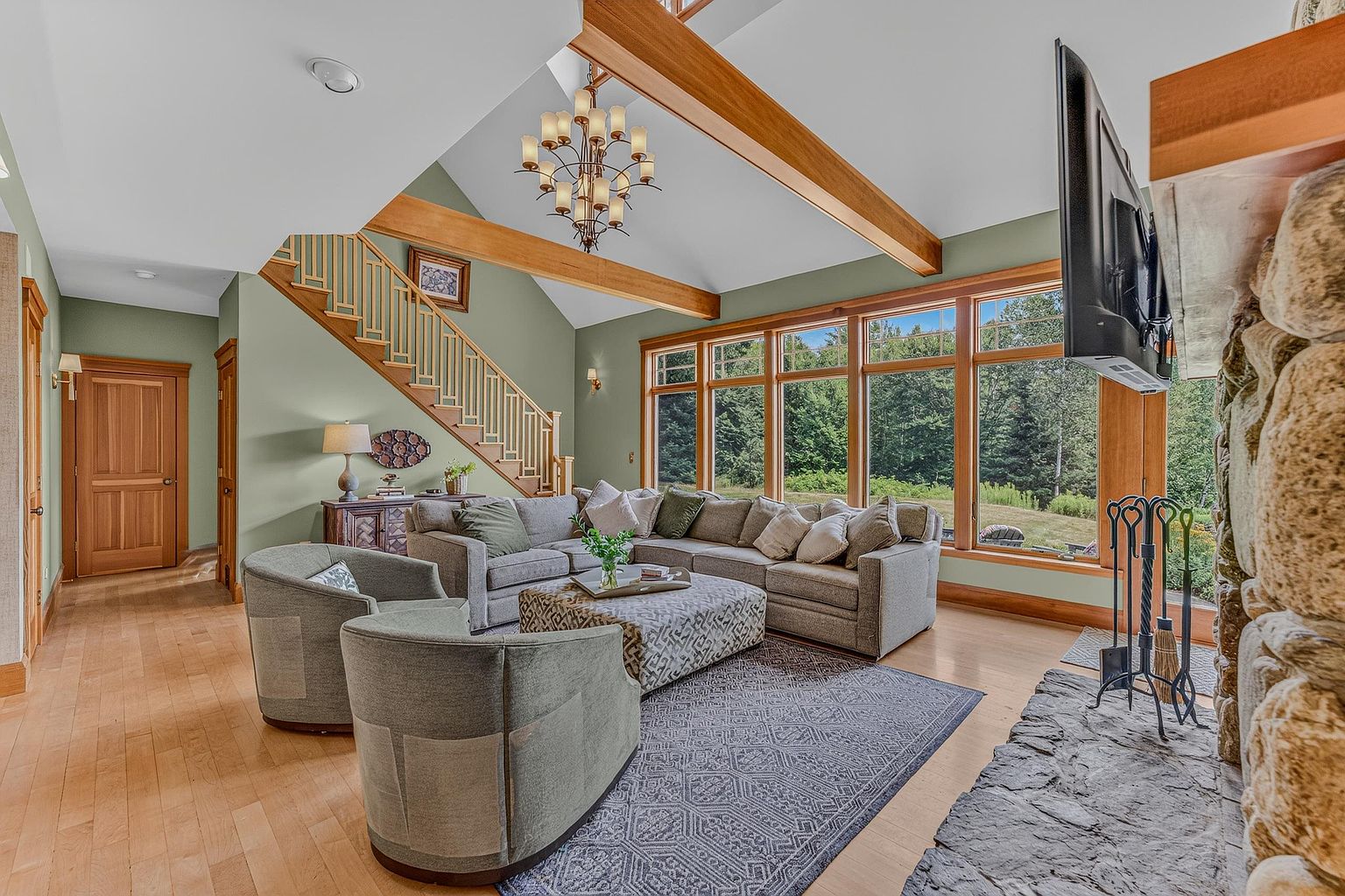 This spacious living room features a vaulted ceiling with exposed wooden beams and a large stone-clad chimney breast. A comfortable sectional sofa and two armchairs are arranged around a patterned ottoman on a large area rug, creating a cozy seating area. Tall windows along the back wall provide abundant natural light and views of the surrounding greenery, while a wooden staircase adds architectural interest to the side of the room.