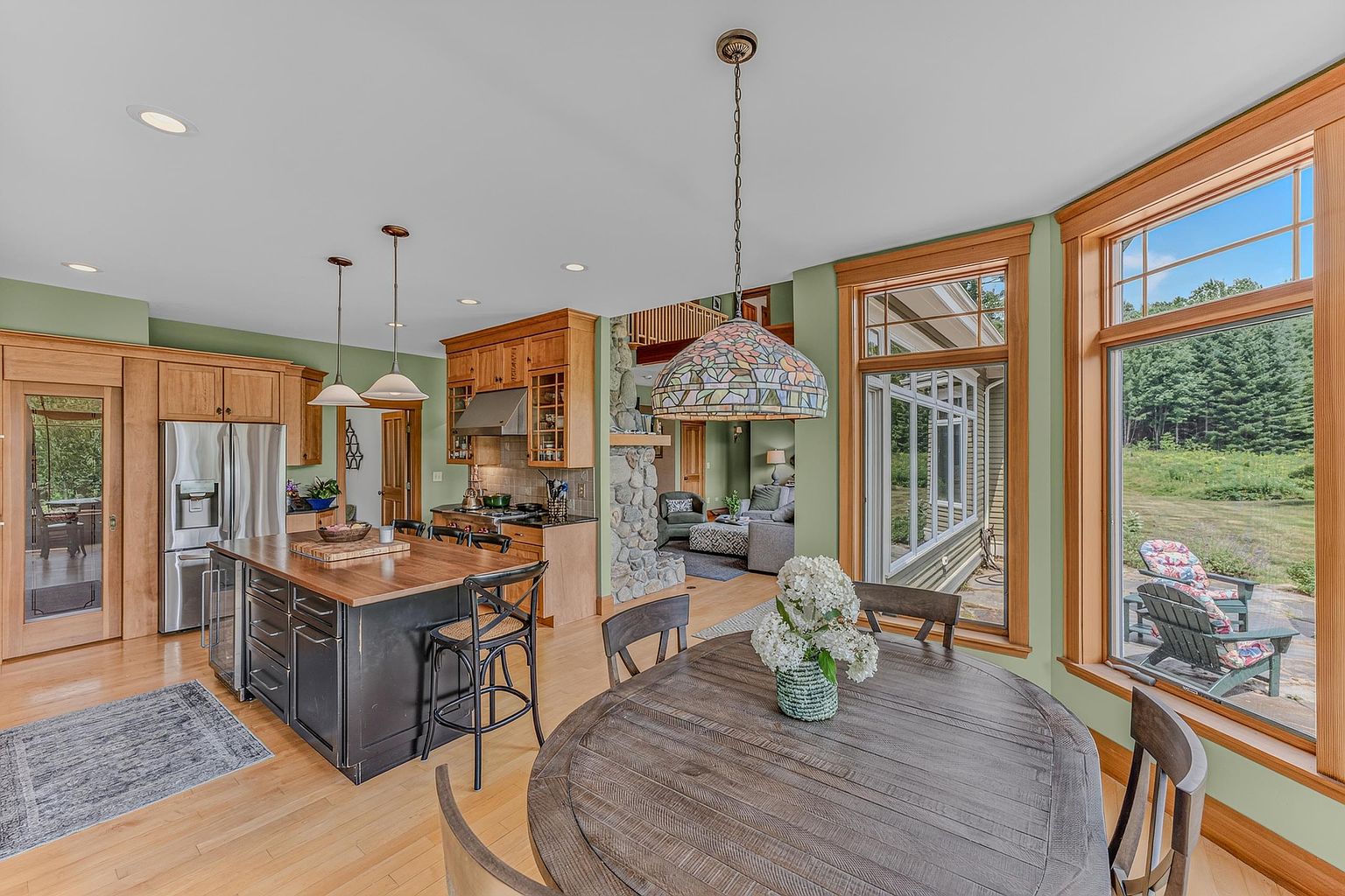 This open-concept kitchen and dining area features a large central island with a wood countertop and dark cabinetry, complemented by light wood flooring. A stained-glass pendant light hangs above a round wooden dining table, while large windows provide abundant natural light and views of the surrounding landscape. The space flows seamlessly into a living area anchored by a prominent stone-clad chimney breast, creating a warm and inviting atmosphere.