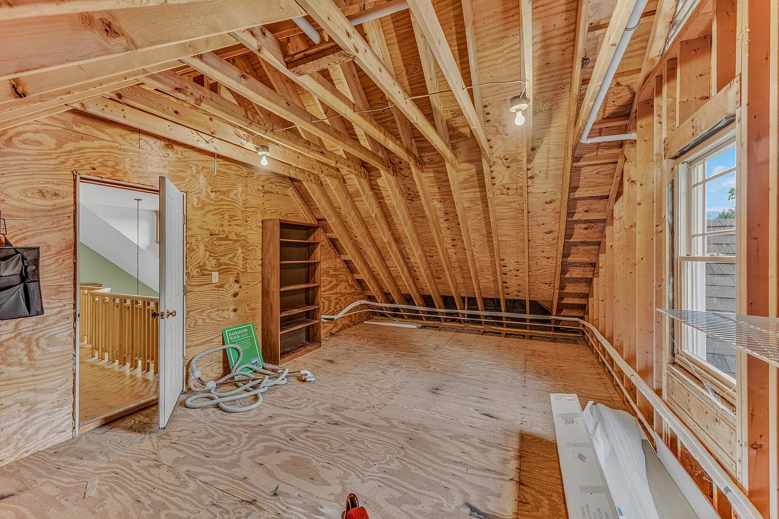 This unfinished attic space features exposed wooden rafters and plywood walls, offering a blank canvas for potential renovation. The room includes a built-in wooden bookshelf, a single window providing natural light, and an open doorway leading to a staircase. The perspective captures the raw, structural character of the space, highlighting its potential for conversion into a functional living area.