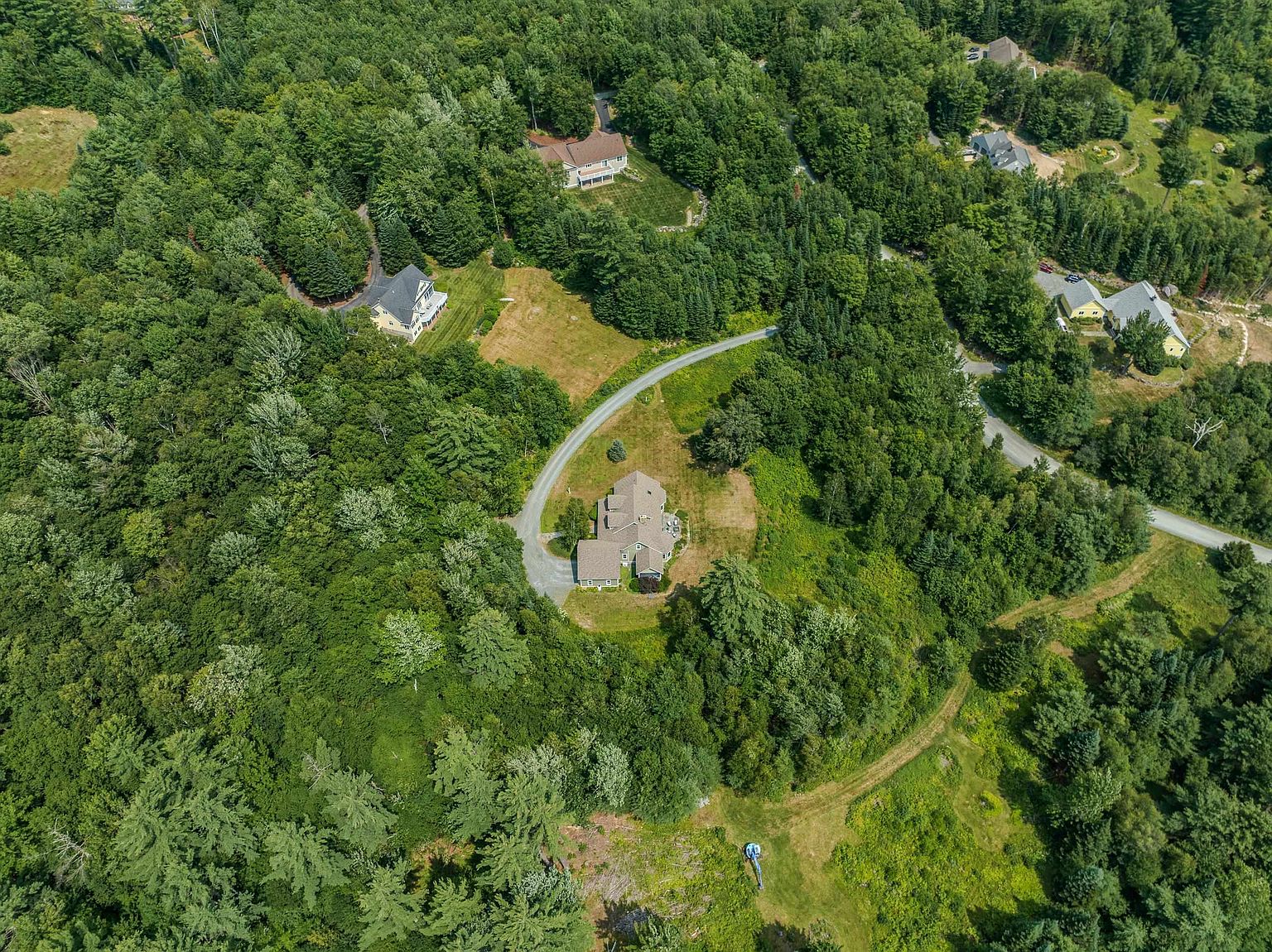This high-angle aerial view captures a secluded residential property nestled within a dense, lush forest. The scene features a primary home with a winding driveway, surrounded by expansive green lawns and several neighboring houses tucked into the wooded landscape. The perspective emphasizes the private, rural setting and the generous amount of surrounding natural greenery.