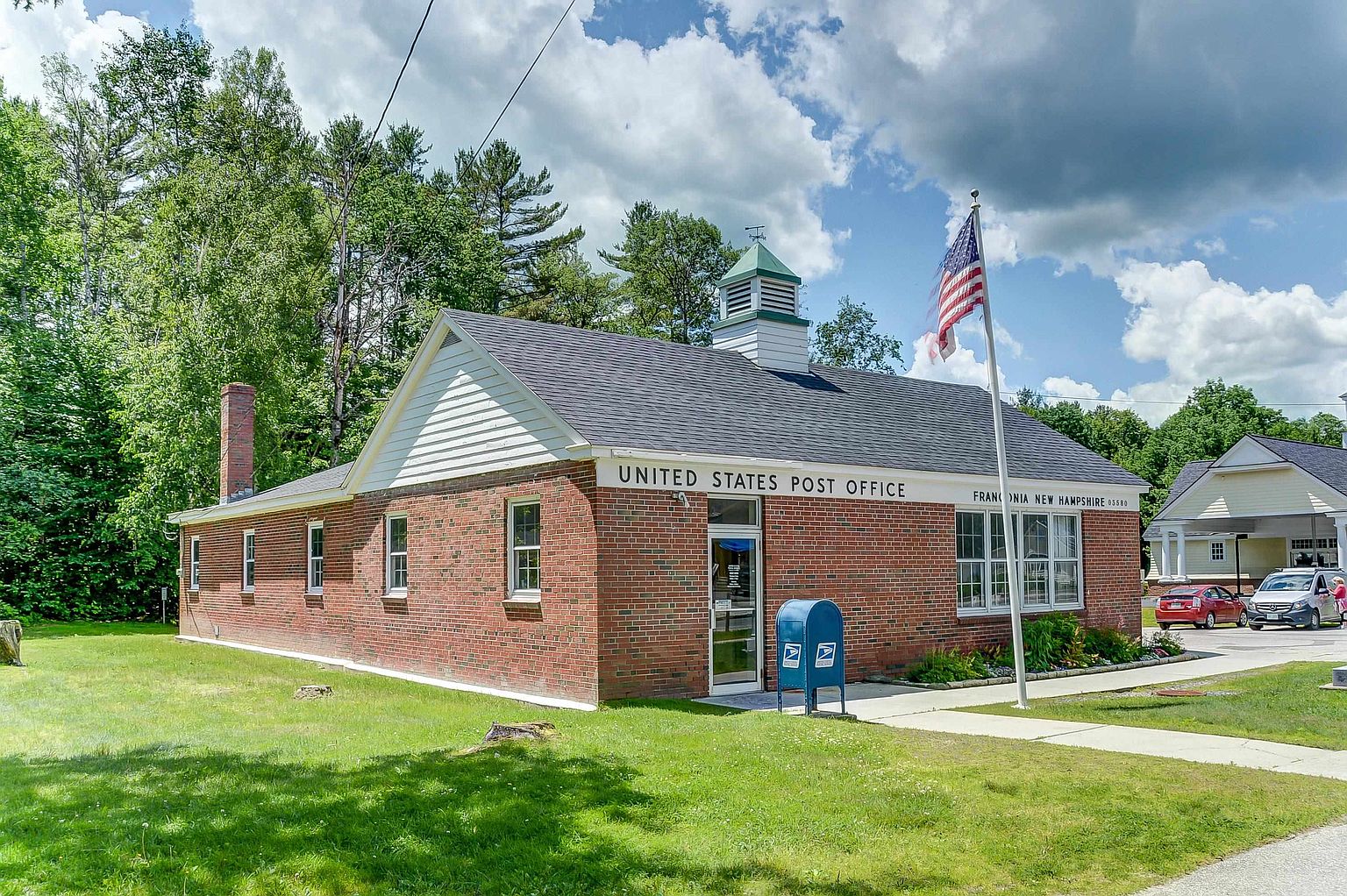 This image captures the front exterior of a classic brick United States Post Office in Franconia, New Hampshire. The building features a traditional gabled roof, a central cupola, and a prominent American flag flying on a pole near the entrance. A blue USPS collection box sits on the concrete walkway, and the surrounding grounds include a well-maintained lawn and mature trees under a bright, partly cloudy sky.