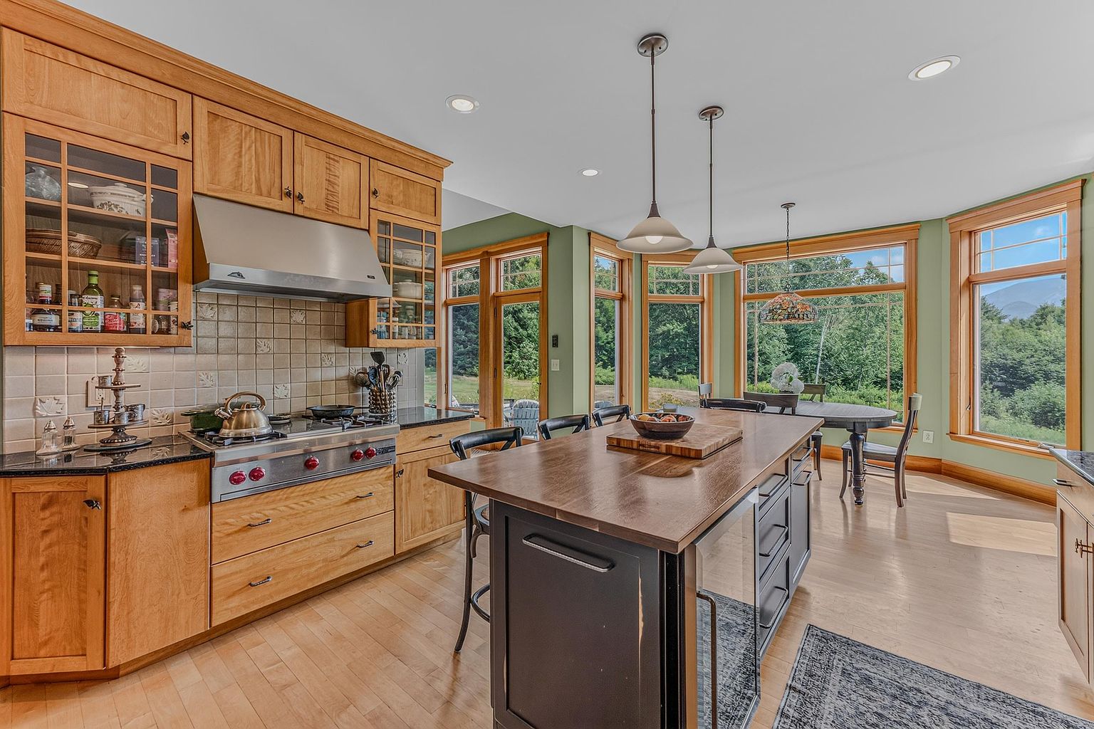 This spacious kitchen features light wood cabinetry, a large central island with a dark base, and a professional-style range with a stainless steel hood. The room is bathed in natural light from a series of large windows that offer expansive views of the surrounding greenery and distant mountains. A dining area is seamlessly integrated into the space, creating an open and inviting atmosphere perfect for entertaining.