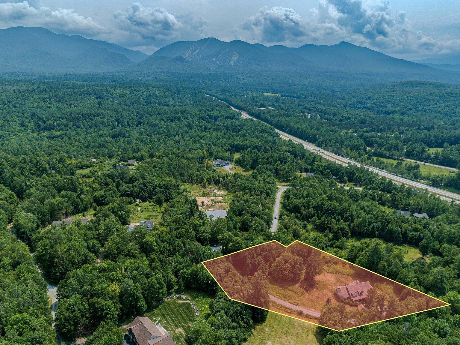 This high-angle aerial view captures a secluded residential property nestled within a lush, forested landscape, with a highlighted boundary overlaying the lot. The scene features a single-story home surrounded by mature trees, a private driveway, and expansive natural grounds, all set against a dramatic backdrop of distant mountain ranges. The perspective emphasizes the property's privacy and its integration into the surrounding rural environment.
