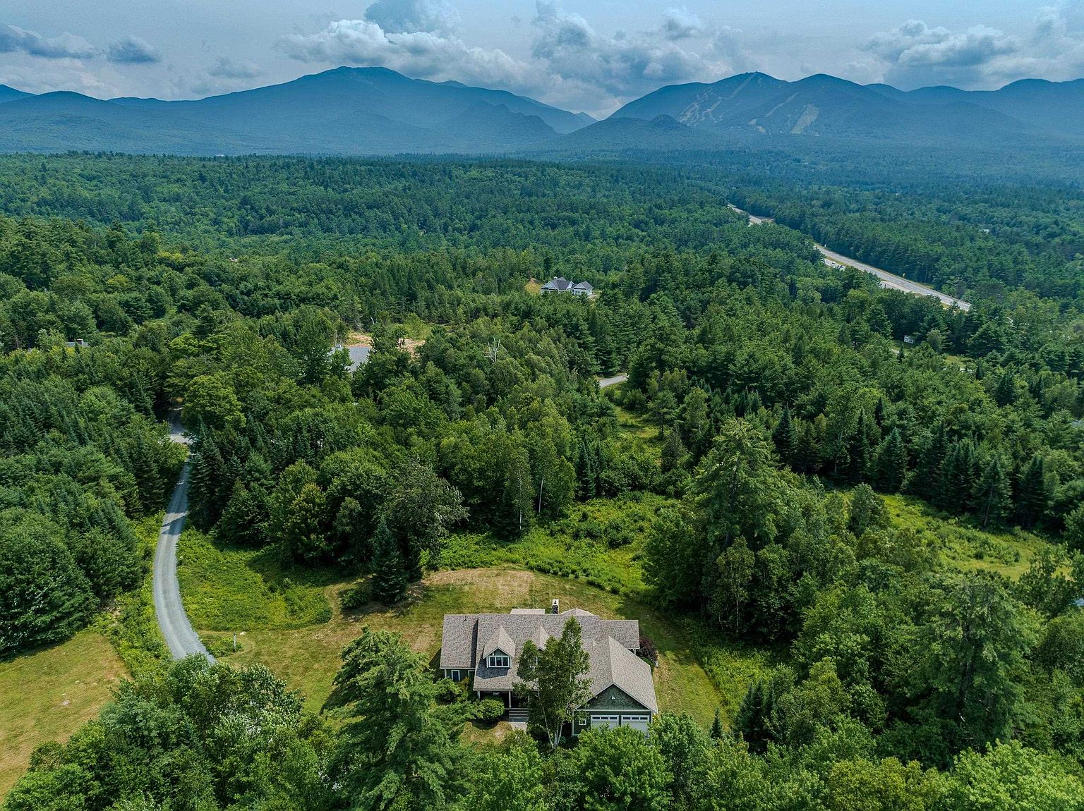 This high-angle aerial shot captures a secluded residential property nestled within a dense, lush forest. The home features a multi-gabled roofline and a neutral-toned exterior, surrounded by expansive greenery and a winding gravel driveway. In the background, majestic mountain ranges rise against a partly cloudy sky, emphasizing the home's serene and private location.