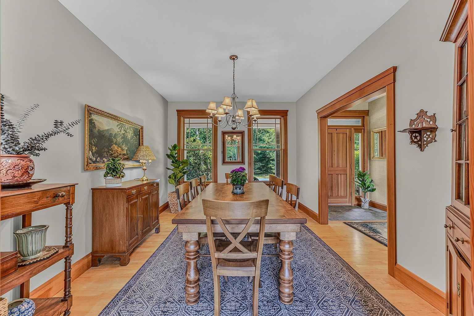 This formal dining room features a large wooden table centered on a patterned blue area rug, surrounded by matching chairs. The space is anchored by a traditional chandelier, a wooden sideboard with a landscape painting above it, and warm wood trim framing the windows and doorways. The room offers a classic, inviting atmosphere with a clear view through to the entryway.