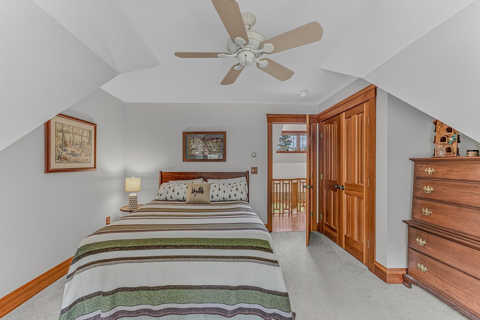 This cozy bedroom features a bed with a striped quilt and patterned pillows, centered against a light-colored wall. A wooden dresser stands to the right, while a ceiling fan provides overhead lighting and air circulation. The room includes a doorway leading to a hallway with a wooden railing, creating an open and inviting atmosphere.