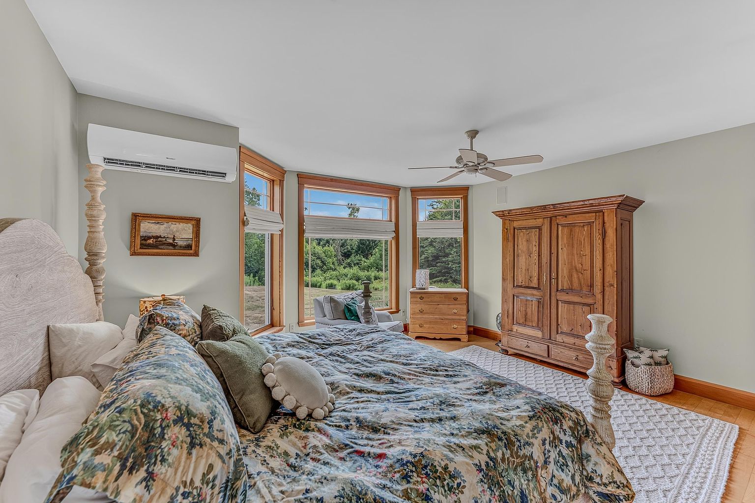 This spacious primary bedroom features a large bed with a patterned floral duvet and a light-colored headboard, positioned centrally in the room. The space is illuminated by natural light streaming through a series of large windows that offer a scenic view of the surrounding greenery. A substantial wooden wardrobe provides ample storage, while a ceiling fan and wall-mounted air conditioning unit ensure climate control, creating a comfortable and serene atmosphere.