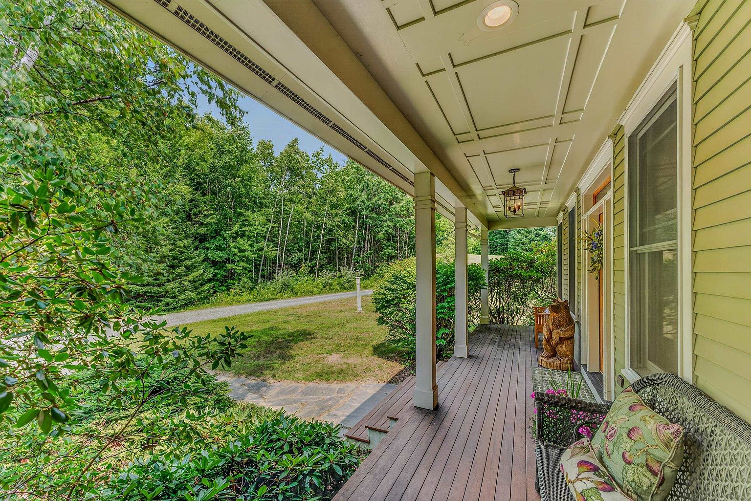 This inviting covered porch features dark wood-toned decking and a light-colored ceiling with recessed lighting and a decorative hanging lantern. A wicker bench with patterned cushions sits near the entrance, complemented by a whimsical wooden bear carving. The porch overlooks a lush, green forest and a gravel driveway, creating a peaceful and secluded atmosphere.