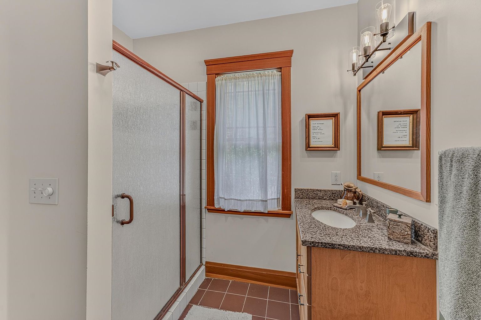 This bathroom features a shower with a textured glass enclosure, a vanity with a stone countertop, and a single sink. A window with a light curtain is framed by wood trim, matching the vanity and mirror frame, while the floor is finished with dark square tiles. The space is illuminated by a multi-light fixture above the mirror, creating a functional and traditional aesthetic.