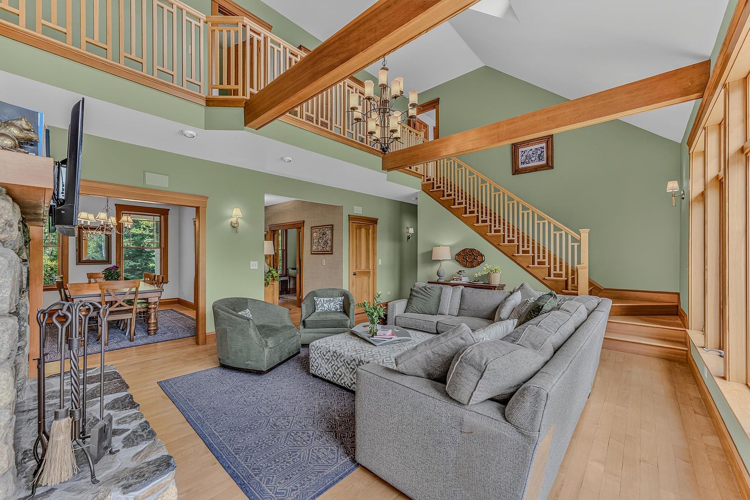 This spacious, open-concept living area features a vaulted ceiling with exposed wooden beams and a prominent stone-clad chimney breast. A large gray sectional sofa and two matching armchairs are arranged around a patterned ottoman on a blue area rug, creating a comfortable seating area. The room flows into a dining space visible through a wide doorway, while a wooden staircase with decorative railings leads to an upper landing, all set against soft green walls and light wood flooring.