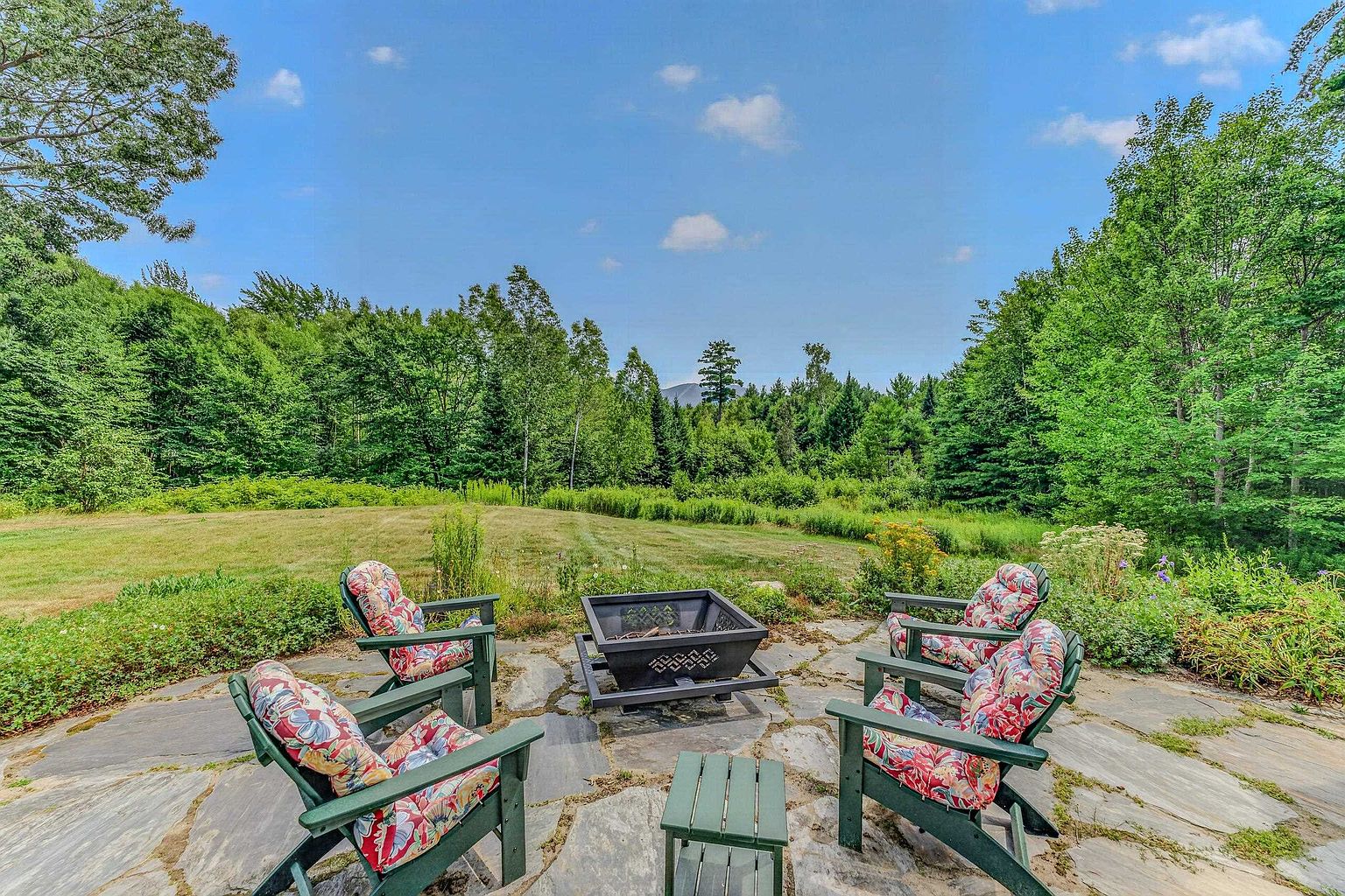 This outdoor living space features a natural stone patio arranged with four green Adirondack chairs and a central metal fire pit. The patio overlooks a lush, expansive grassy lawn that transitions into a dense forest backdrop under a bright blue sky. The setting offers a serene and private atmosphere, perfect for relaxation or entertaining in a natural environment.