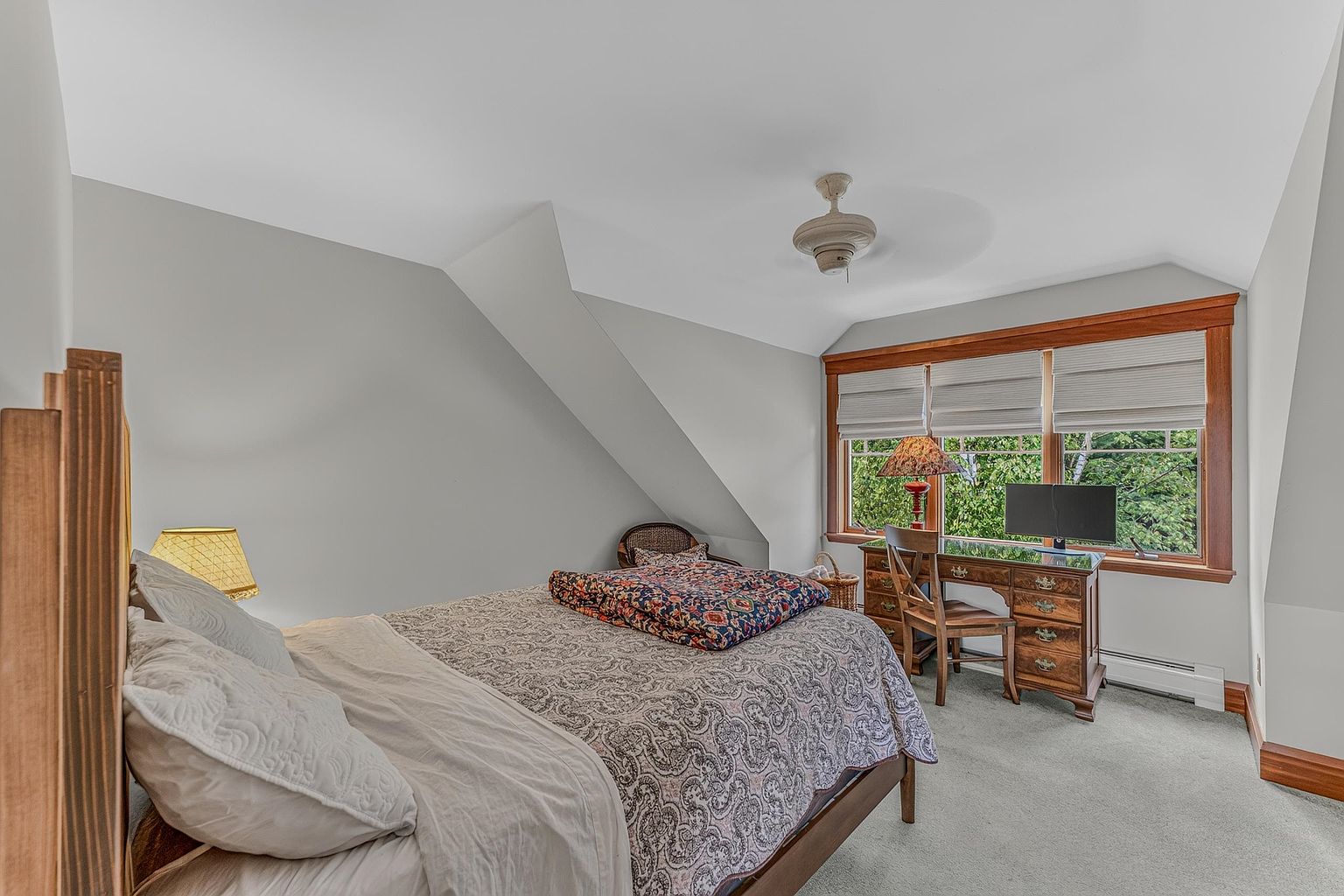 This cozy bedroom features a bed with a patterned quilt and a wooden headboard, positioned under a sloped ceiling. A wooden desk with a computer monitor and a chair is tucked into the corner near a window with light-filtering shades. The room is finished with neutral wall paint and light-colored carpeting, creating a comfortable and functional living space.